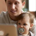 Man kijkt op een tablet met een baby die een speen met een blauw randje draagt.