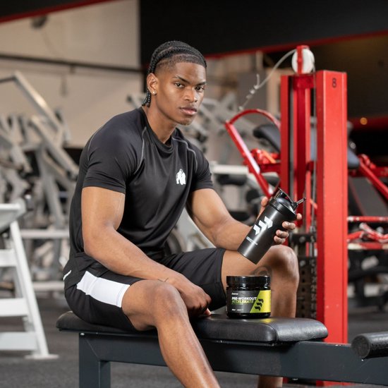 Man met donkere huid, in zwart sportshirt, houdt zwarte shaker en "Empose Pre-Workout Tropical" pot vast in fitnessruimte.