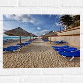 WallClassics - Muursticker - Blauwe Ligbedden op Strand met Rieten Parasols - 40x30 cm Foto op Muursticker