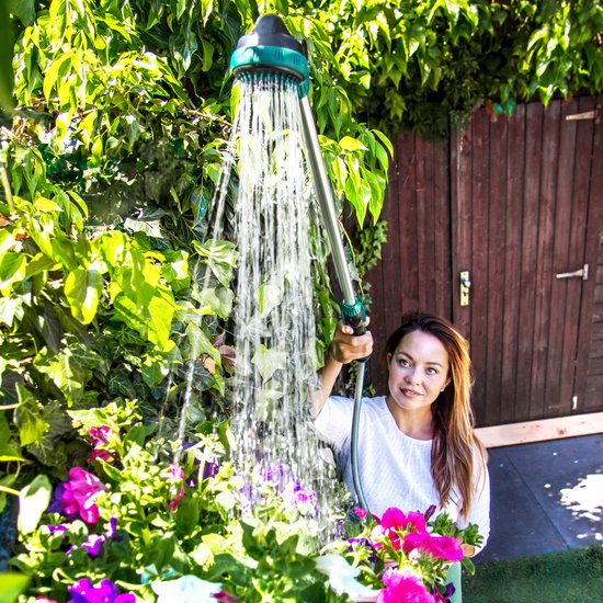 Een vrouw in een witte top sproeit met een lange zilveren sproeilans met groene accenten, bloemen en groen op de achtergrond.