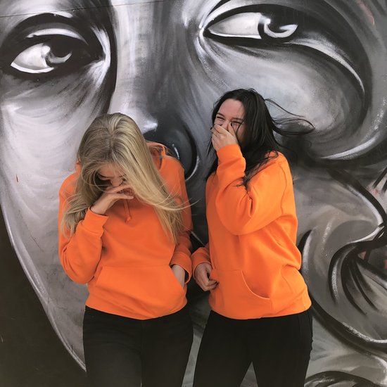 Deux femmes riant, adossées à une fresque murale, portant toutes deux un sweat à capuche orange vif.