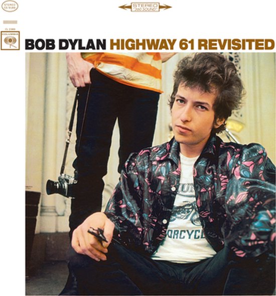 Bob Dylan met een zwart-roze panterprint jasje, een wit T-shirt met "Motorcycle" erop, en zwarte broek.