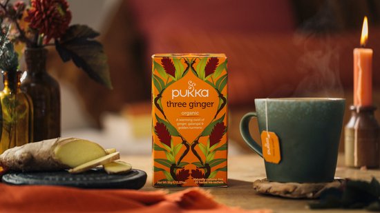 Pukka three ginger thee verpakking naast een mok dampende thee, gember en een kaars.
