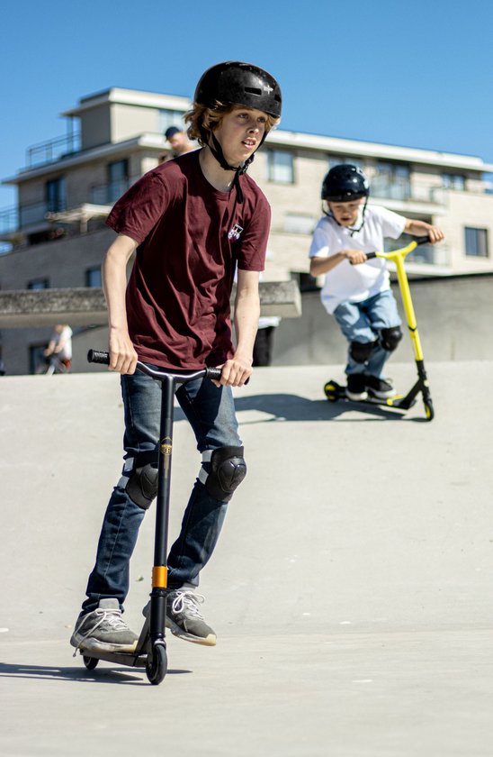 Jongen in donkerrode shirt, blauwe jeans en kniebeschermers rijdt op zwarte step.