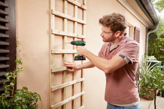 Man boort met groene Bosch accuboormachine in een houten klimplantenrek.