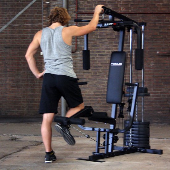 Man met krullend haar, grijs T-shirt en zwarte korte broek oefent benen aan het Focus Fitness Unit 2 krachtstation.