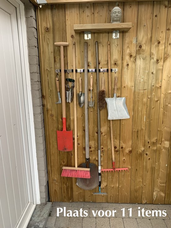 Houten wand met diverse tuingereedschappen, waaronder bezems, schoppen en een hark, opgehangen aan haken.