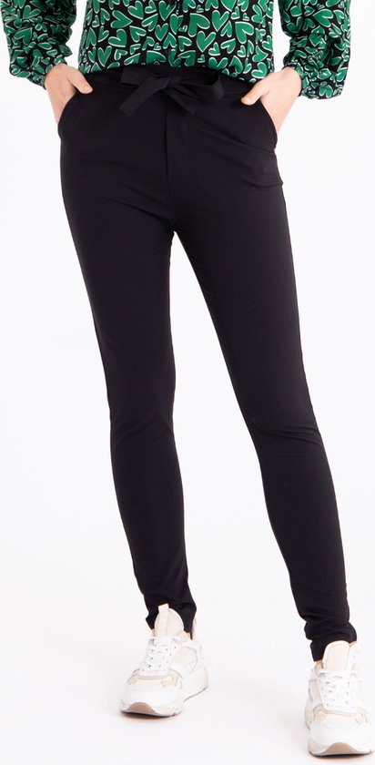 Een vrouw in een zwarte broek met strikceintuur en witte sneakers.