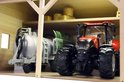 Close-up van een rode tractor in een houten landbouwloods, naast een grijze tank met aanhanger.
