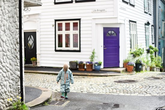 Kind in een lichtgroen regenpak met visgraatpatroon, lopend in een straat met kasseien en witte huizen.