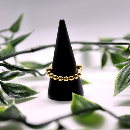 Gouden ring met balletjes op een zwarte ringhouder, omringd door groene bladeren.