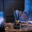 Zwarte glazen diffuser van Ted Sparks met gouden dop en zwarte stokjes, op een marmeren tafel.