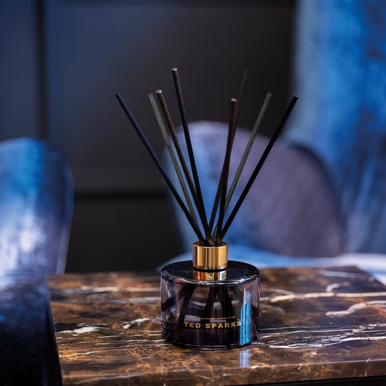 Zwarte glazen diffuser van Ted Sparks met gouden dop en zwarte stokjes, op een marmeren tafel.