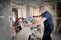 Man met bril, donkerblauwe polo, zaagt houten balk met Metabo afkortzaag KGS254M.