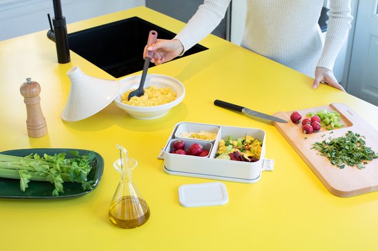 Dans une cuisine avec un plan de travail jaune, une boîte à pain grise remplie de salade et de fruits. Des ingrédients sont préparés.