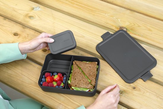 Handen plaatsen een deksel op een donkergrijze bento lunchbox met sandwich, fruit en salade.