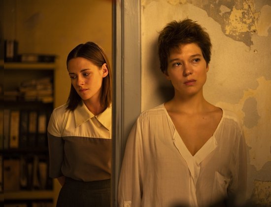 Kristen Stewart en Léa Seydoux staan dichtbij elkaar bij een deurpost. Achtergrond met boekenplanken.