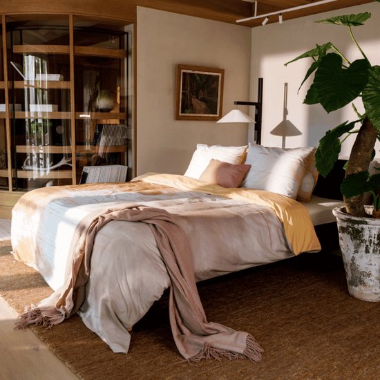 Een slaapkamer met een tweepersoonsbed opgemaakt met dekbedovertrek met een abstract patroon in oranje en wit tinten. Een beige plaid ligt over het bed.