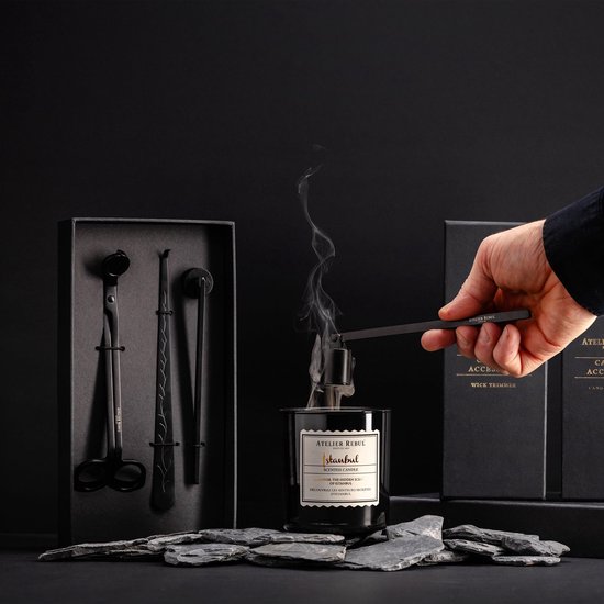 Een hand gebruikt Atelier Rebul wick trimmer om een brandende Atelier Rebul Istanbul geurende kaars te doven. Kaart is donker glas met een label.