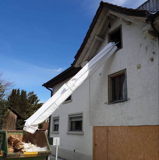 Een witte stortkoker hangt uit een raam en loopt naar een container met puin.
