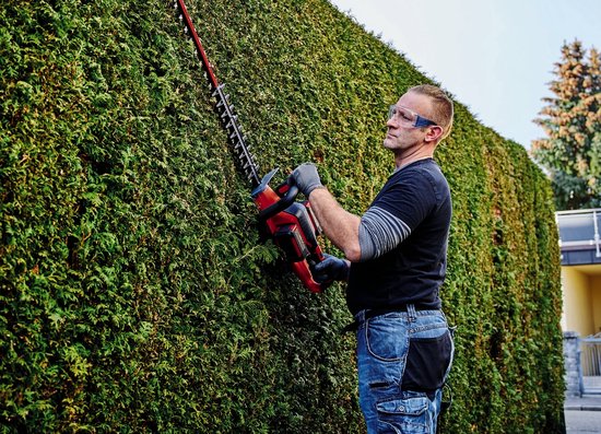Man snoeit een groene heg met een rode en zwarte Einhell heggenschaar.