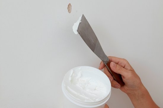 Hand met spatel en pot witte houtrotvuller, repareert gat in witte muur.