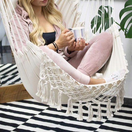 Vrouw in lichte roze outfit zit in witte hangstoel met macramé en franjes, houdt mok vast.
