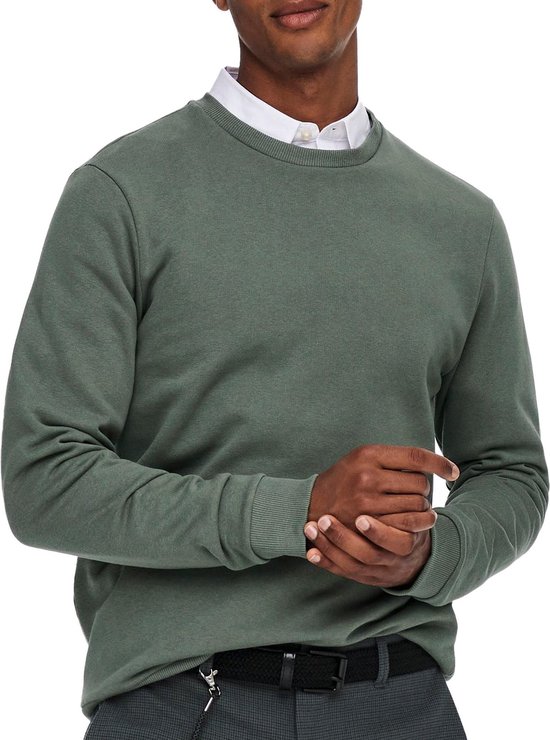 Man draagt een groene trui met ronde hals over een wit overhemd. Hij vouwt zijn handen.
