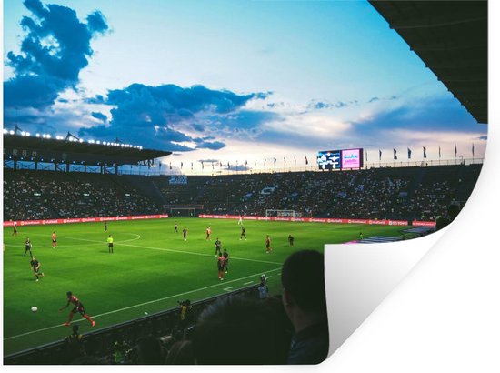 Stade de football avec public et joueurs sur pelouse verte au crépuscule, avec des nuages dans le ciel bleu.
