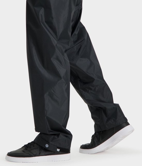 Model draagt een zwarte, glanzende regenbroek met witte sneakers.