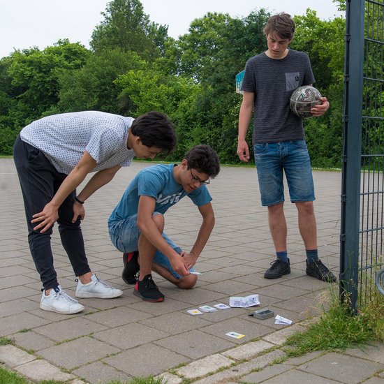 Drie jongens op een plein, gebogen over kaarten. Een jongen met bal op de achtergrond.