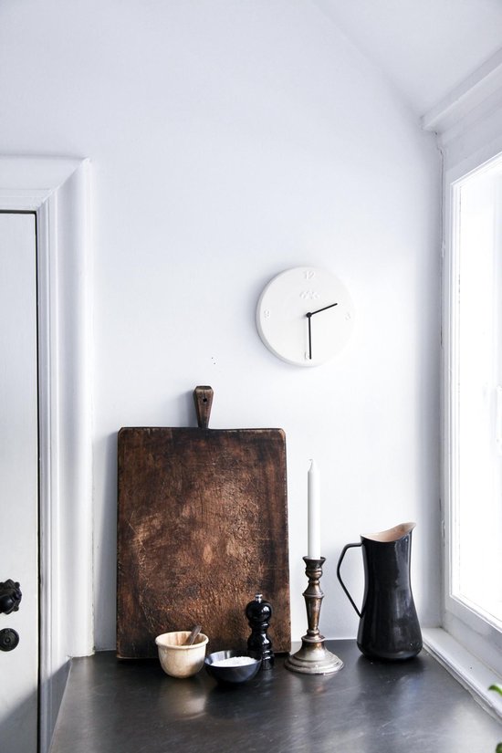 Een witte klok met zwarte wijzers hangt boven een oud houten snijplank op een aanrecht.