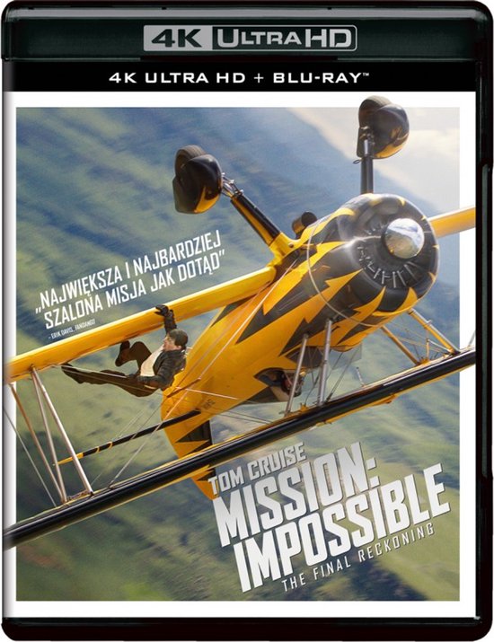 Tom Cruise hangt aan een gele dubbeldekker met zwarte strepen, vliegend in een groene, heuvelachtige omgeving.