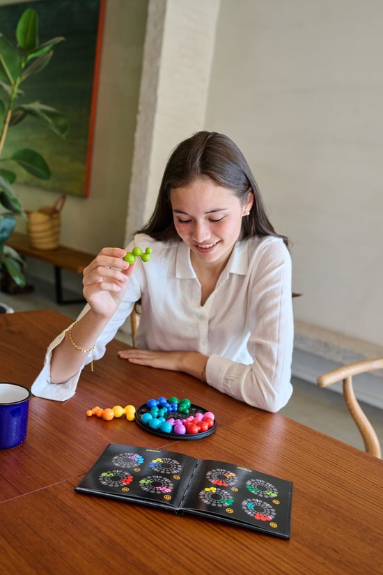 Een jong meisje met een witte blouse glimlacht terwijl ze het IQ Circle spel speelt aan een houten tafel. Ze houdt een groen stuk vast.