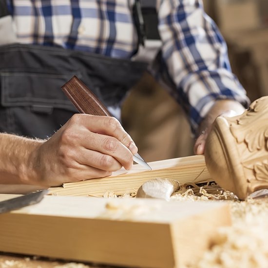 Hand met houtsnijmes van donker hout, die houtsnijdt. Houtsnippers op de tafel.