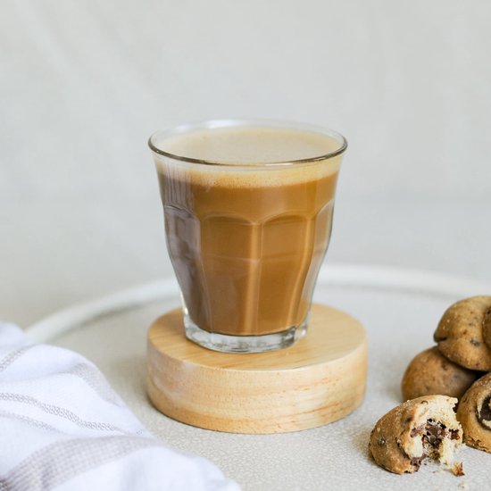 Un verre de café avec de la mousse, sur un sous-verre en bois. À côté de biscuits et d'un tissu.