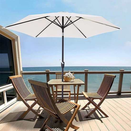 Een witte parasol staat open boven een houten tafel en stoelen op een terras aan zee.