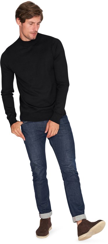 Man buigt voorover, handen in zakken, draagt zwarte coltrui en jeans.