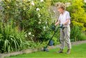 Vrouw in witte blouse en beige broek maait met groene grastrimmer in een tuin met witte bloemen.