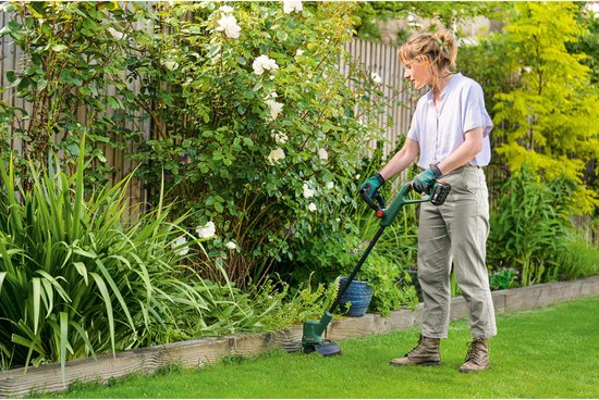 Vrouw in witte blouse en beige broek maait met groene grastrimmer in een tuin met witte bloemen.