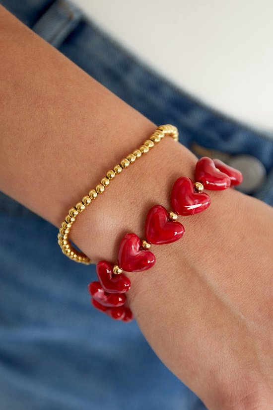 Gouden kraaltjesarmband met rode hartvormige bedeltjes, gedragen om een pols.