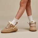 Kind met beige UGG sneakers aan, witte sokken met ruches en een wit T-shirt.