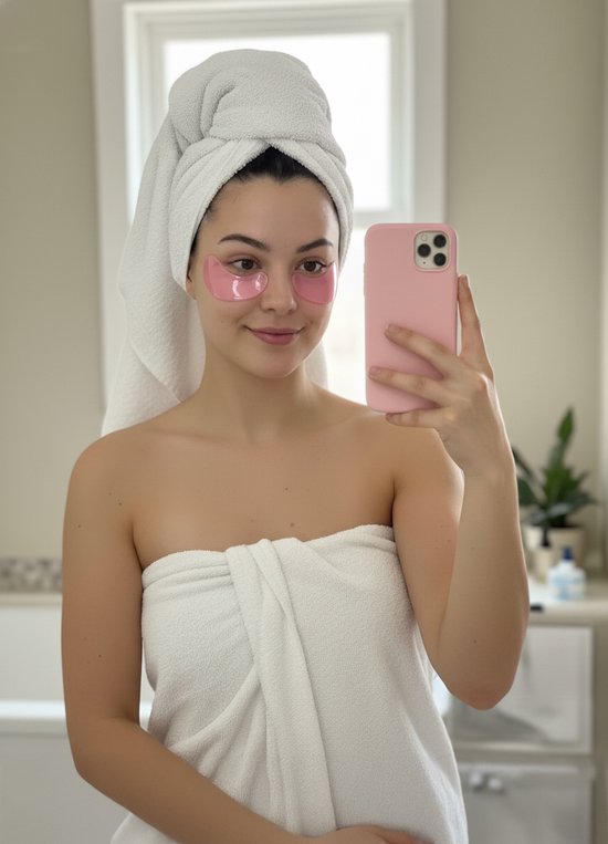 Een vrouw met een witte handdoek om haar hoofd en lichaam, draagt roze oogpatches en houdt een roze telefoon vast.