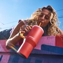 Vrouw met krullend haar drinkt uit een koraalrode Kambukka beker met lichtgeel handvat en lichtroze deksel, buiten op een roze muur.