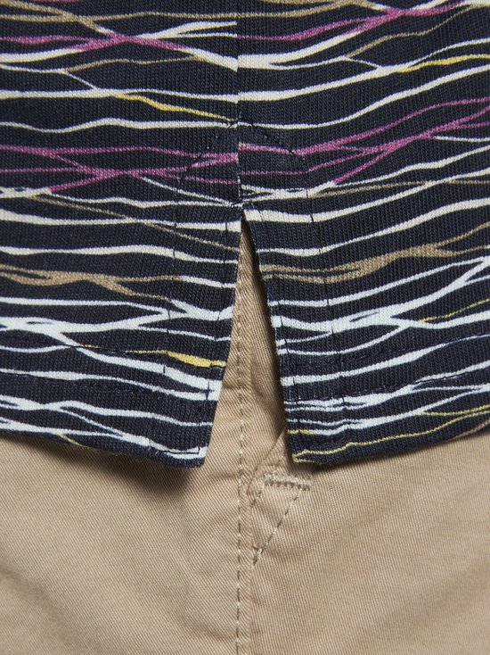 Detail van de zoom van een donkerblauwe polo met golvend streepjespatroon in paars, geel en wit, over een beige broek.