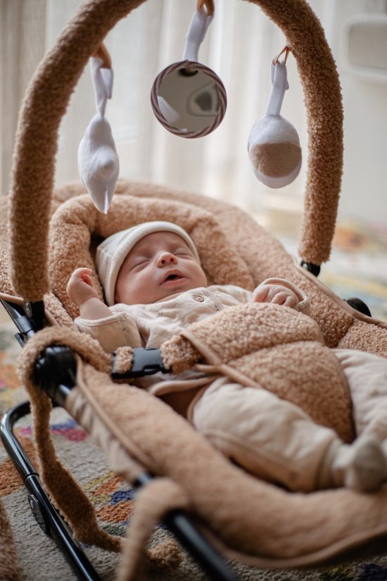 Baby in beige outfit en mutsje slaapt in een bruine wipstoel met speelboog.