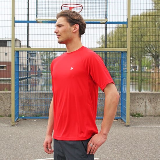 Man staat op een sportveld, draagt een rood sportshirt met korte mouwen en een logo.