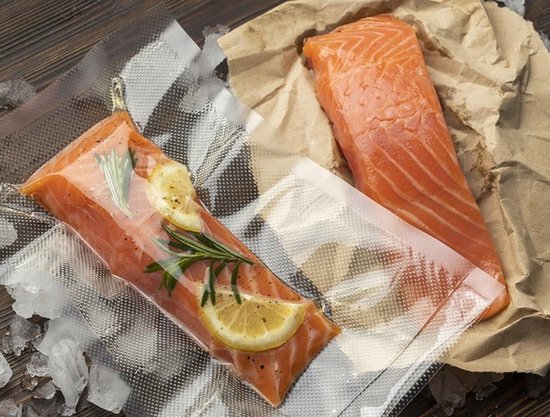 Un filet de saumon dans un sac sous vide avec du citron et du romarin, à côté d'un morceau de saumon sur du papier brun.