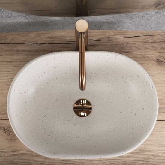 Ovaal beige waskom met spikkels en koperkleurige kraan op houten badmeubel.