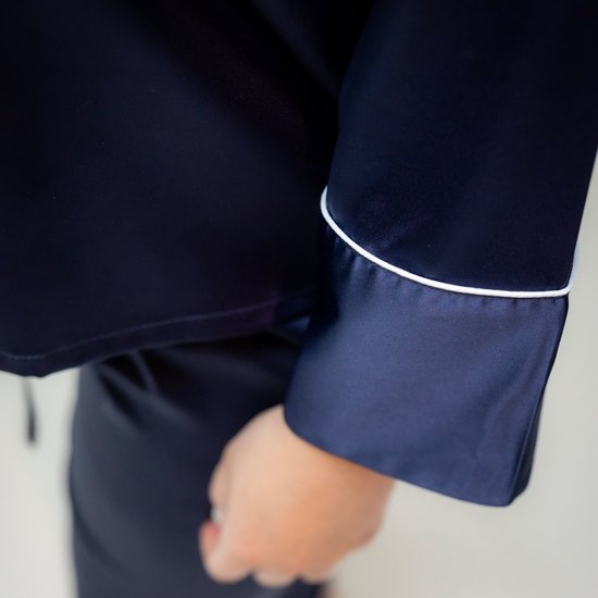 Close-up van de mouw van een donkerblauwe satijnen pyjamajas met witte biesjes en de hand van het model.